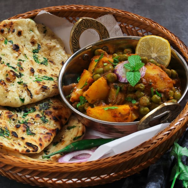 Aloo Matar, an Indian potato and pea curry, served in a metal bowl with naan bread in a woven basket.