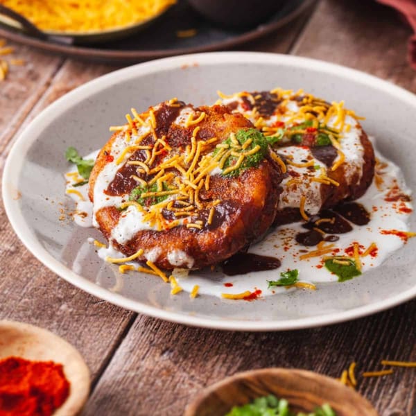 Two golden-brown Indian aloo tikki patties topped with yogurt, chutneys, and sev on a plate.